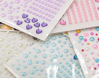 May include: Assortment of self-adhesive pearl embellishments in various colours and sizes. The pearls are arranged on sheets with a clear plastic backing. The colours include white, light blue, pink, and purple.