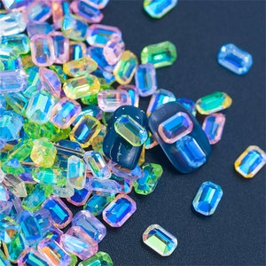 May include: Assortment of iridescent, faceted, rectangular gems in shades of green, blue, pink, and yellow. The gems are scattered on a dark blue surface.