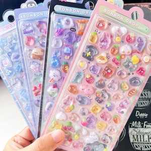 May include: Four packs of colourful, 3D Bonbon Drop stickers. Each pack features a variety of cute, cartoon-like designs, including animals, food items, and musical notes. The stickers are in shades of pink, purple, blue, and orange.