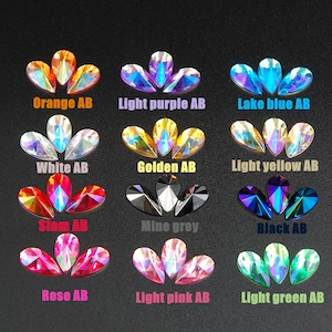 Peut inclure: Une collection de strass irisés en forme de goutte d'eau, de différentes couleurs : orange, violet clair, bleu lac, blanc, doré, jaune clair, siam, gris mine, noir, rose, rose clair et vert clair. Chaque groupe est étiqueté avec son nom de couleur et "AB".