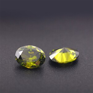 May include: Two green oval-shaped gemstones with a faceted cut. The gemstones are on a dark gray surface.