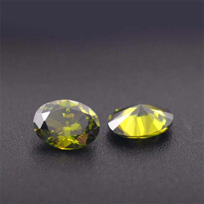 May include: Two green oval-shaped gemstones with a faceted cut. The gemstones are on a dark gray surface.