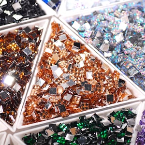 Può includere: Assortimento di piccoli strass quadrati iridescenti in vari colori, tra cui nero, verde, blu e marrone, esposti in contenitori di plastica bianca.