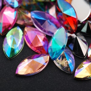 May include: Assortment of iridescent, faceted, marquise-shaped rhinestones in various colours, including pink, blue, green, and gold. The rhinestones are scattered on a black surface.