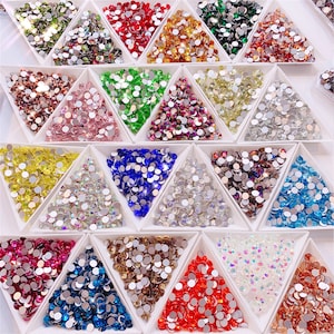 May include: Assortment of colorful rhinestones in various shapes and sizes, displayed in triangular compartments on a white surface.