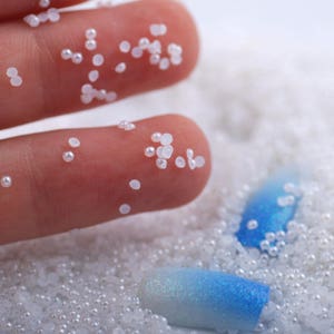 May include: White pearl beads scattered on a surface with two blue and white nail tips. The beads are small and round, and the nail tips are long and oval shaped.