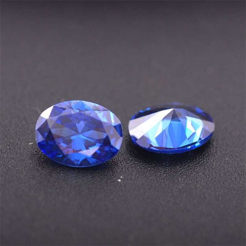 May include: Two oval-shaped, faceted, blue gemstones on a black surface.