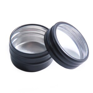 May include: Two black metal tins with a clear lid. The tin on the left is open and shows a silver interior. The tin on the right has a clear lid.
