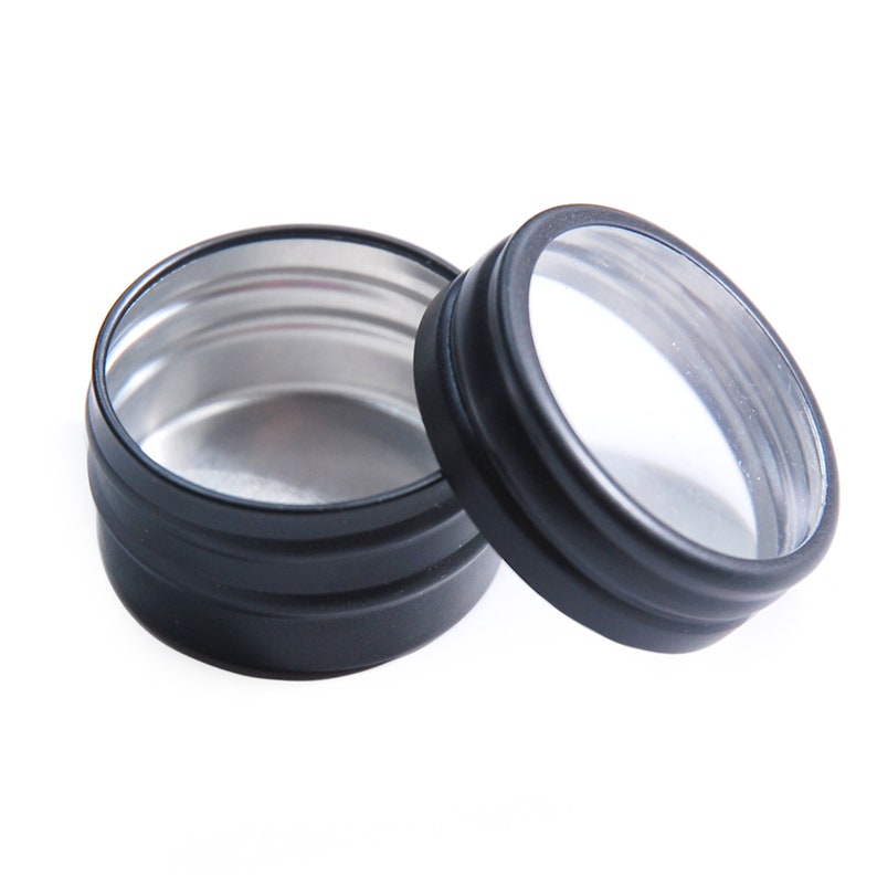 May include: Two black metal tins with a clear lid. The tin on the left is open and shows a silver interior. The tin on the right has a clear lid.