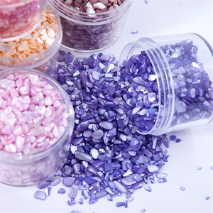 May include: Close-up of several clear plastic jars filled with colorful, iridescent shell chips. The colors include shades of purple, pink, and peach. One jar is tipped over, spilling the purple chips onto a white surface.