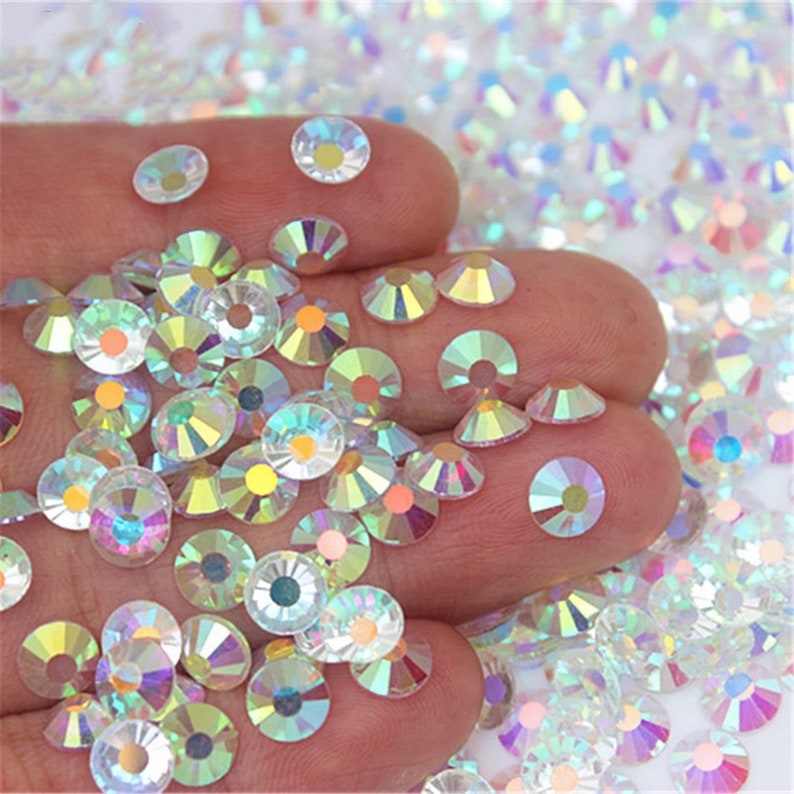 May include: A hand holding a large quantity of iridescent clear rhinestones. The rhinestones are various sizes and shapes, with a rainbow effect.