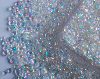 May include: A close-up of iridescent white glass beads, scattered on a light blue surface. The beads are small and oval-shaped, with a smooth, glossy finish.