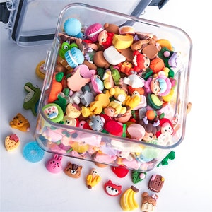 May include: A clear plastic container filled with colorful, miniature food and animal-themed charms. The charms include bananas, strawberries, bears, and macarons. A few charms are scattered outside the container, including a green alligator and a blue donut.