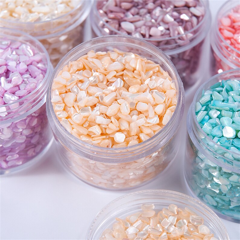 May include: A collection of small, iridescent, shell-like pieces in various colors, including peach, pink, purple, and turquoise, are displayed in clear plastic jars. The pieces have a natural, irregular shape and a pearlescent sheen.