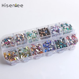 Puede incluir: Un contenedor de plástico transparente con 12 compartimentos llenos de diferentes colores de piedras de imitación ovaladas. Las piedras de imitación son iridiscentes y tienen un efecto arcoíris.