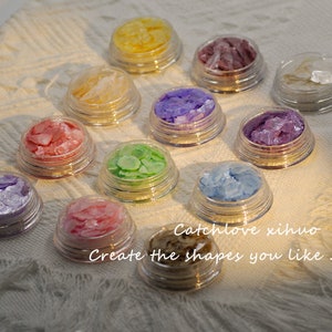 May include: Ten small jars filled with iridescent, colourful, and glittery flakes. The jars are arranged in a semi-circle on a white surface. The text "Catchlove xihua - Create the shapes you like" is visible below the jars.