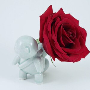 May include: A light blue ceramic figurine holds a large, vibrant red rose. The figurine has a rounded body and small limbs. The rose is in full bloom, with many petals. The background is plain white.