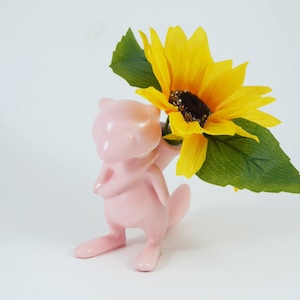May include: A pink ceramic figurine of a creature holding a yellow sunflower with green leaves. The figurine has a smooth, glossy finish and is set against a white background. The sunflower has a dark brown center.