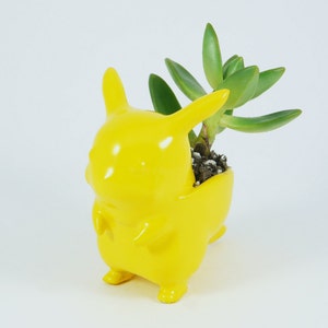May include: A bright yellow Pikachu-shaped ceramic planter holding a green succulent plant. The planter has a glossy finish and features Pikachu's iconic ears, cheeks, and tail. The succulent has thick, green leaves and is planted in soil.
