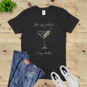 Op de afbeelding: Zwart T-shirt met een wit martini-glas en olijven, en de tekst "You say potato I say Vodka".