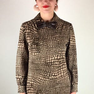 May include: A brown and beige dress with a crocodile print pattern. The dress has a long sleeve and a bow at the collar. The model is wearing black lace-up shoes.