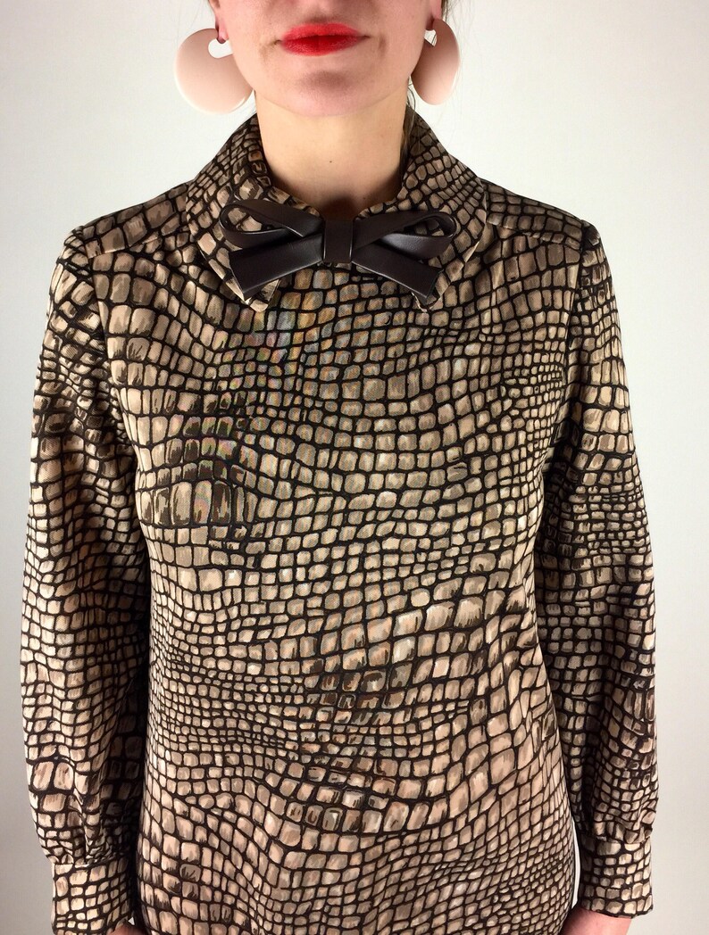 May include: A brown and beige long-sleeved blouse with a faux crocodile skin pattern. The blouse has a high collar and a brown bow tie.