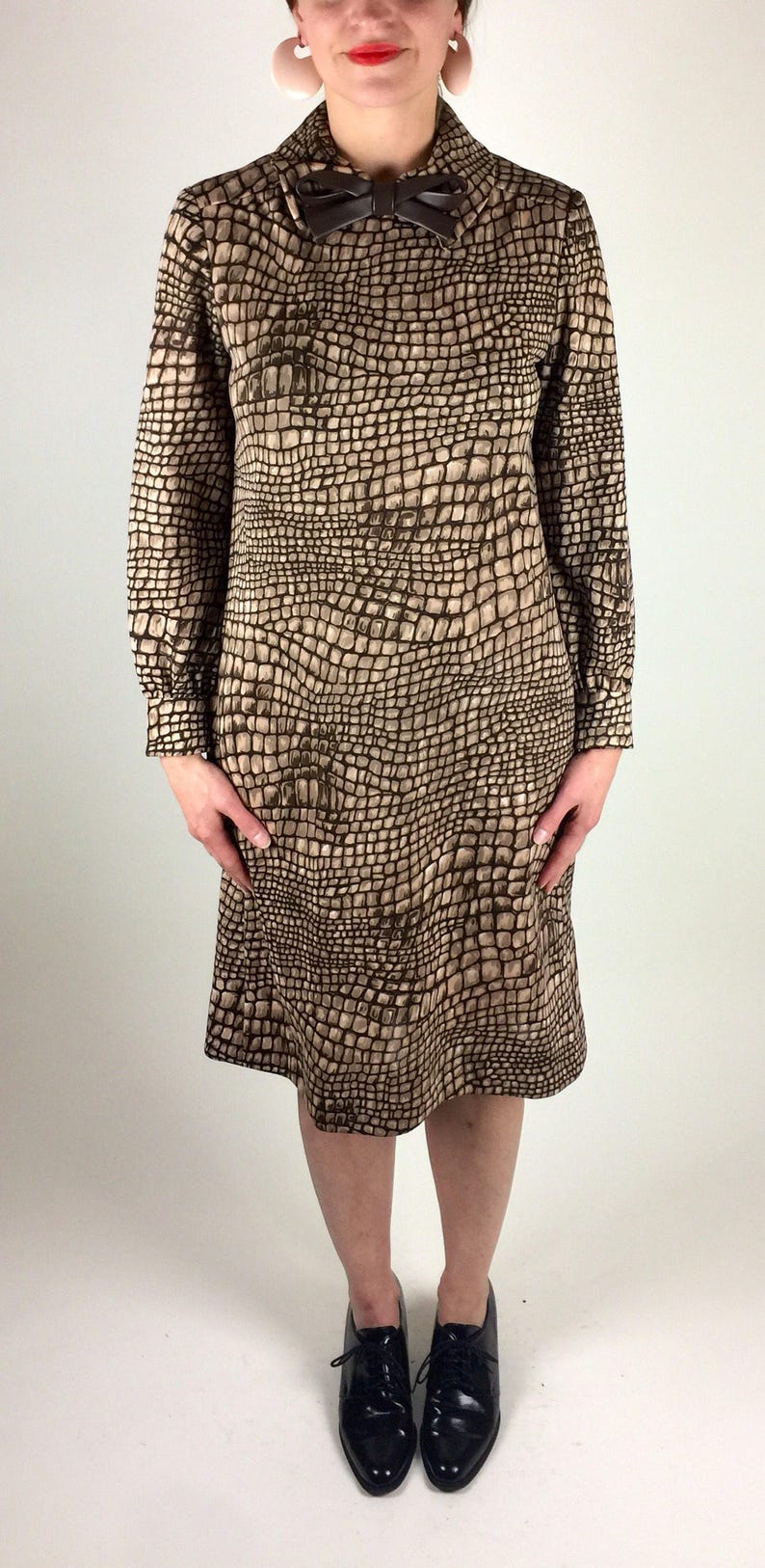 May include: A brown and beige dress with a crocodile print pattern. The dress has a long sleeve and a bow at the collar. The model is wearing black lace-up shoes.