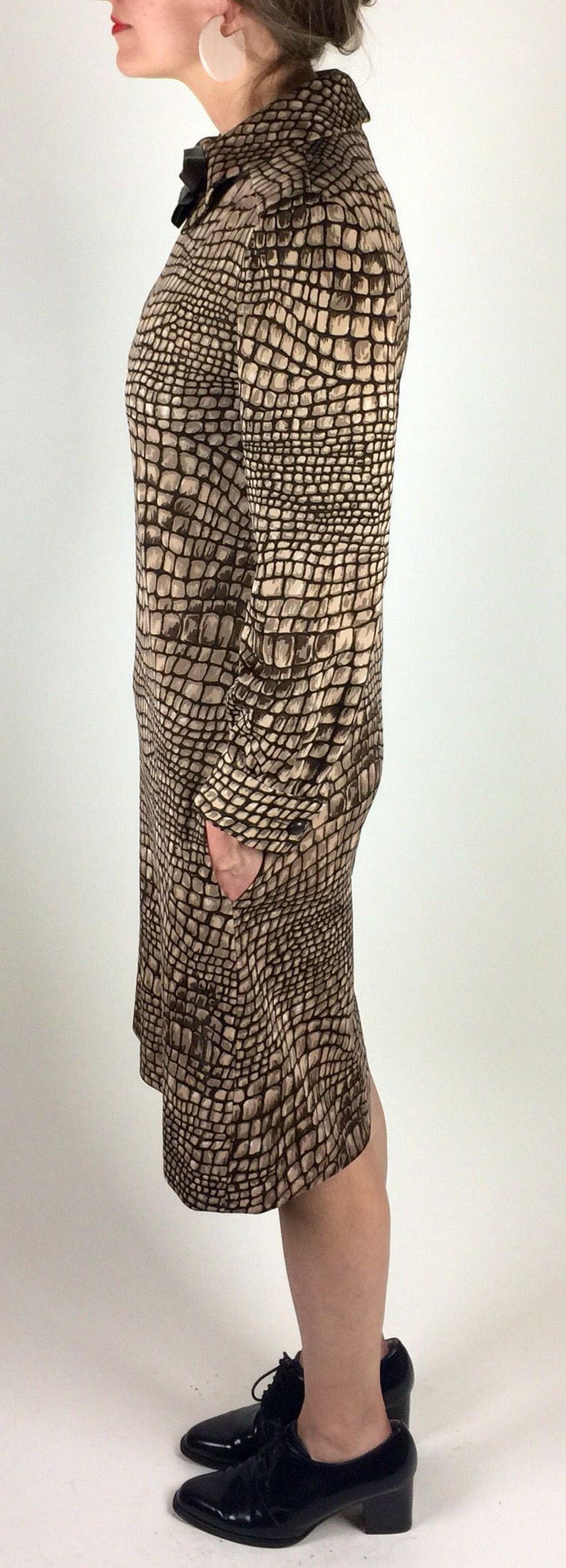 May include: A brown and beige dress with a crocodile print pattern. The dress has long sleeves and a collared neckline. The model is wearing black heeled shoes.