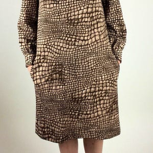 May include: A brown and beige dress with a crocodile print pattern. The dress has a bow at the neckline and long sleeves. The model is wearing black lace-up shoes.