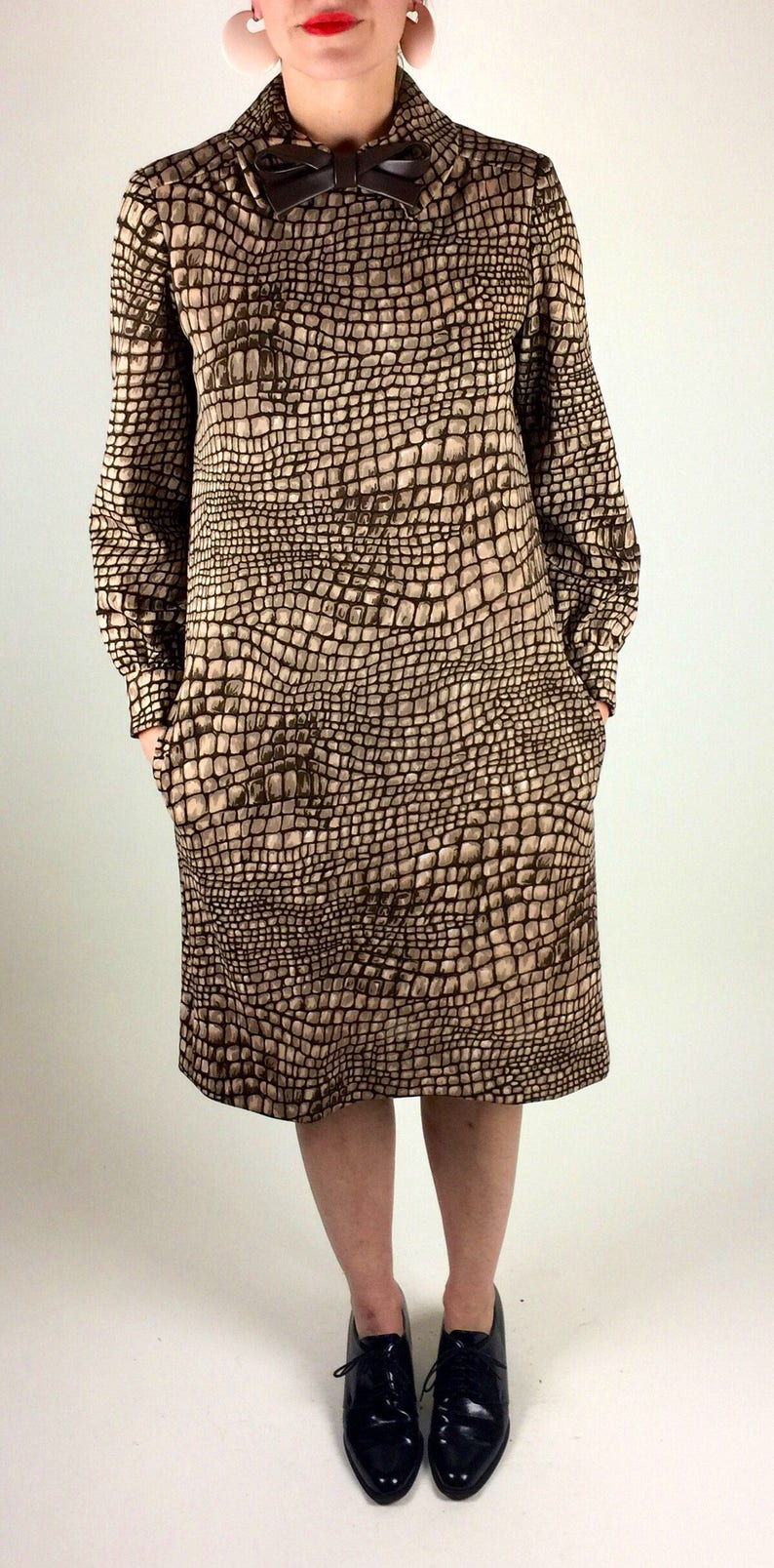 May include: A brown and beige dress with a crocodile print pattern. The dress has a bow at the neckline and long sleeves. The model is wearing black lace-up shoes.
