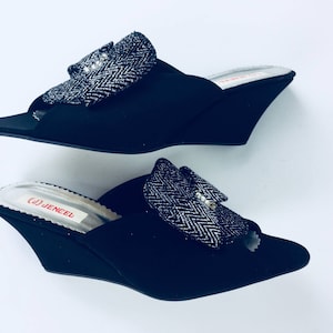 May include: A pair of black wedge sandals with a silver and black patterned bow on the front. The sandals have a red label that says "GJ JENCEL".