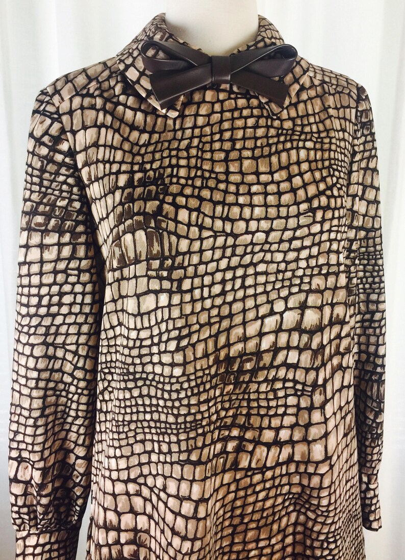 May include: A brown and beige long-sleeved dress with a crocodile print pattern. The dress has a brown faux leather bow at the collar.