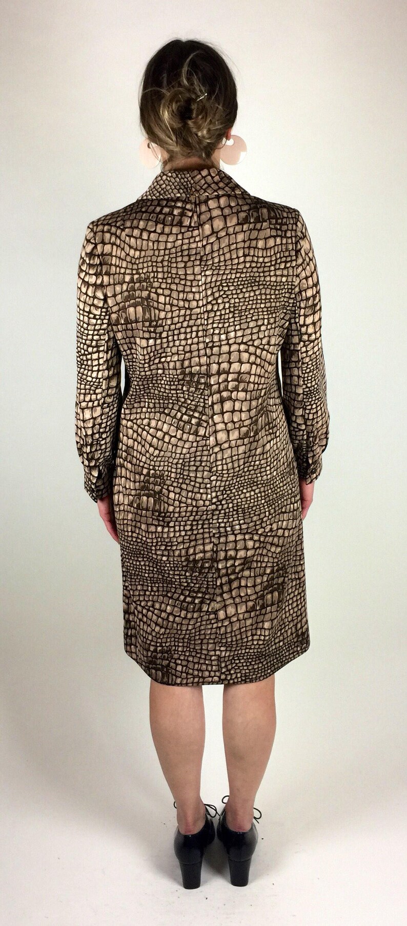 May include: A brown and beige dress with a crocodile print pattern. The dress has long sleeves and a knee-length skirt. The model is wearing black heeled shoes.