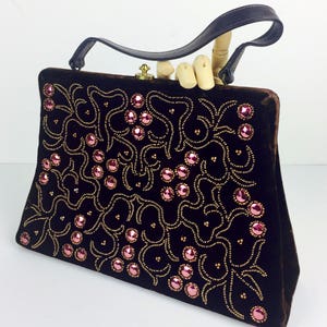 May include: A dark brown velvet handbag with a gold clasp and a black leather handle. The bag is decorated with a floral pattern of gold beads and pink rhinestones.