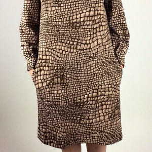 May include: A brown and beige dress with a crocodile print pattern. The dress has a bow at the collar and long sleeves. The model is wearing black lace-up shoes.