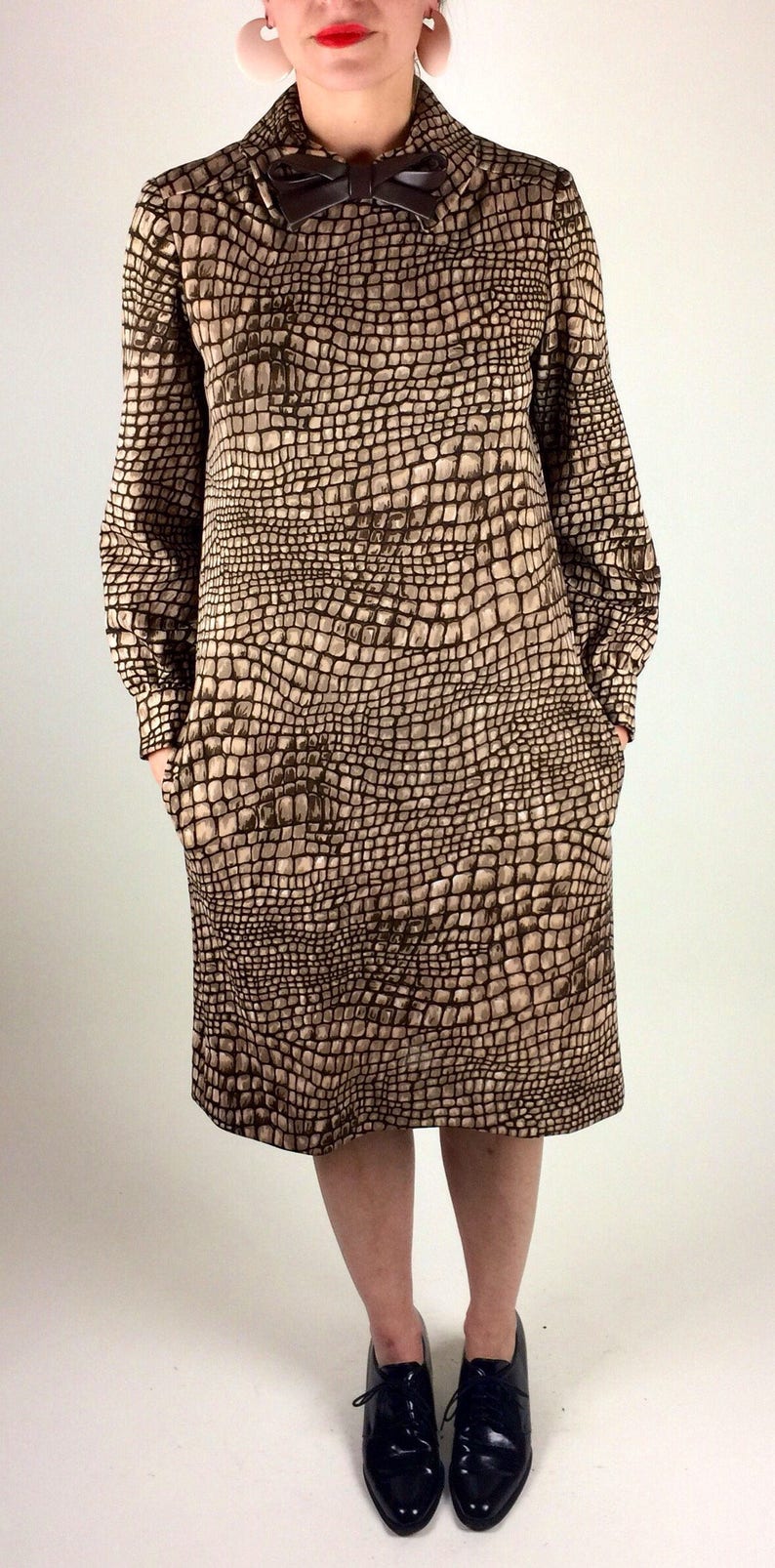 May include: A brown and beige dress with a crocodile print pattern. The dress has a bow at the collar and long sleeves. The model is wearing black lace-up shoes.