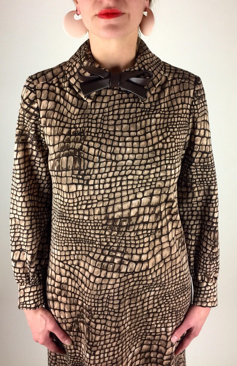May include: A brown and beige dress with a crocodile print pattern. The dress has a long sleeve and a bow at the collar.