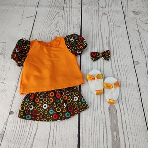 May include: A doll outfit with a bright orange top, a brown skirt with colorful circles, a white headband with a brown bow, and a pair of white sandals with orange straps.