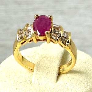 May include: A gold ring featuring a large oval ruby center stone. The ring is flanked by baguette-cut diamonds on each side. The band is a polished gold. The ring is displayed on a white surface.