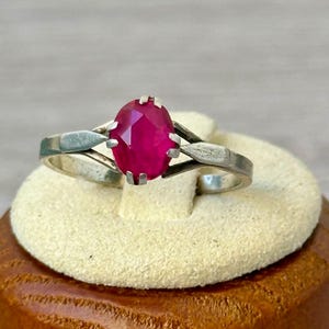 May include: A silver ring featuring an oval, faceted, ruby-red gemstone. The ring's band has a split design with decorative elements on the sides. The gemstone is set in a prong setting, showcasing its vibrant color and shape.