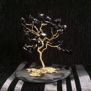 Op de afbeelding: Een gouden draadboom sculptuur met zwarte edelsteen bladeren. De boom wordt getoond op een zwarte leisteen basis.