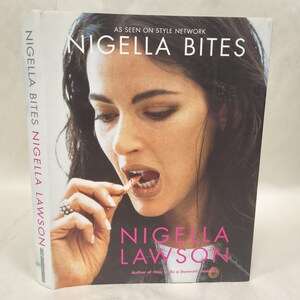Könnte beinhalten: Ein gebundenes Buch mit dem Titel Nigella Bites von Nigella Lawson. Das Cover zeigt ein Foto einer Person, die ein Stück Essen isst, mit dem Text AS SEEN ON STYLE NETWORK, NIGELLA BITES und NIGELLA LAWSON in weißer und rosa Schrift.