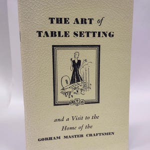 May include: A vintage book titled "THE ART of TABLE SETTING" with a cream-colored cover. The cover features a black and white illustration of a woman setting a table. The text "and a Visit to the Home of the GORHAM MASTER CRAFTSMEN" is also visible.