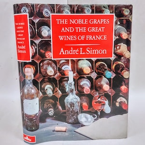 May include: A book cover featuring a wine cellar with numerous bottles. The title "THE NOBLE GRAPES AND THE GREAT WINES OF FRANCE" is displayed in red. The author's name, André L. Simon, is also visible. A wine glass and bottle are in the foreground.