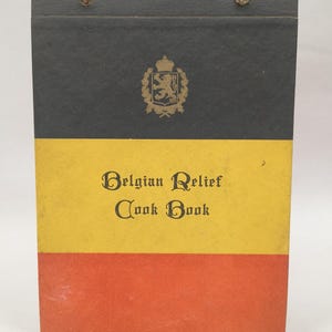 May include: A vertical rectangular cookbook bound with two metal rings at the top. The cover features horizontal black, yellow, and red stripes. A gold emblem of a lion is centred on the black stripe, and the text "Belgian Relief Cook Book" is printed in black on the yellow stripe.