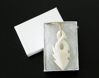 May include: A white bone pendant carved in the shape of a fish hook, with intricate details, is displayed in a white box with a white background. The pendant is on a beige cord.