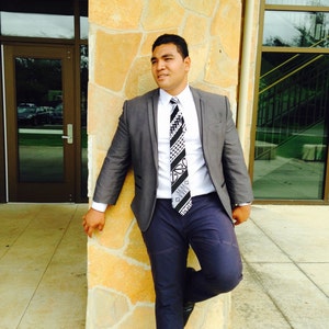 May include: A man wearing a gray suit jacket with a white shirt and a black and white patterned tie. He is standing in front of a stone wall.