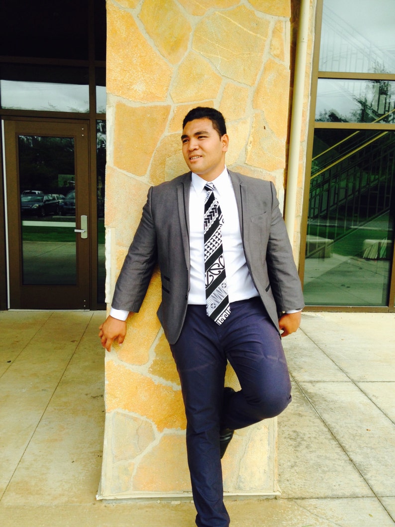 May include: A man wearing a gray suit jacket with a white shirt and a black and white patterned tie. He is standing in front of a stone wall.