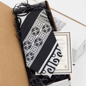 May include: A black and white patterned tie in a cardboard box with black shredded paper. The tie has a geometric design with a white border. The box has a white label with text about the tie.