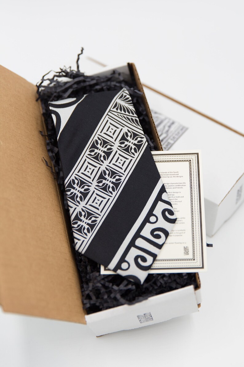 May include: A black and white patterned tie in a cardboard box with black shredded paper. The tie has a geometric design with a white border. The box has a white label with text about the tie.